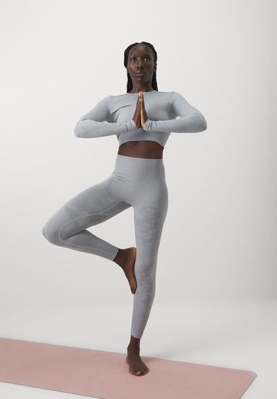 Un crop top și leggings atletici de culoare gri deschis, cu o textură netedă. Figura stă pe un picior pe un covoraș de yoga roz, cu mâinile într-o poziție de rugăciune.