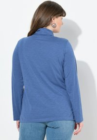 Modrý top s dlouhým rukávem a vysokým límcem, vyrobený z texturované látky. Spodní lem je rovný a je kombinován se světle modrými džínami.