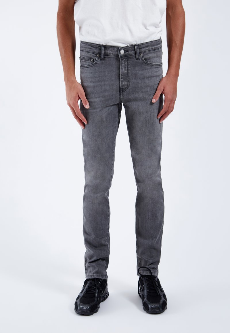 Jeans de mezclilla gris de corte ajustado, con cinco bolsillos, trabillas para el cinturón y un ligero acabado desgastado. Combinados con zapatillas deportivas negras.