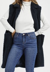 Czarna, fakturowana kamizelka z kołnierzem typu szal, noszona na białym golfie, w połączeniu z niebieskimi dżinsami z wysokim stanem. Widoczne kieszenie na kamizelce.