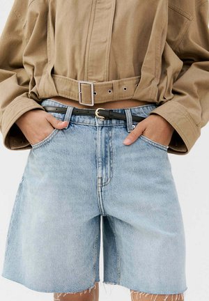 Osoba nosiaca svetlomodré džínsové krátke nohavice s vysokým pásom, čiernym opaskom a krátku béžovú bundu, ruky vreckách krátkych nohavíc.