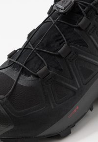 Chaussure de sport noire avec une tige en mesh, des détails texturés, un système de laçage élastique et un logo Contagrip sur le côté.