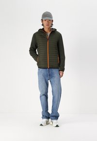 Olivgrüne Pufferjacke mit orangen Akzenten, aus leichtem Stoff, mit Kapuze, kombiniert mit hellblauen Jeans und weißen Sneakers.