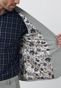 Giacca da abito grigio chiaro con fodera in tessuto floreale nei toni del blu e del crema. Camicia con motivo a scacchi blu navy e linee sottili.