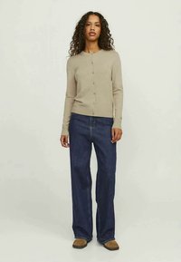 Beige gebreide cardigan met lange mouwen en een knopenfront, gecombineerd met donkerblauwe wijde jeans. Het model staat tegen een neutrale achtergrond.