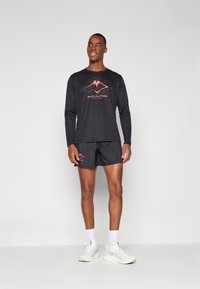 T-shirt de sport à manches longues noir avec logo orange, associé à un short noir arborant un logo similaire, porté avec des baskets et des chaussettes blanches.