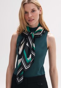 Zijden sjaal met geometrische patronen in zwart, groen en room, elegant om de hals geknoopt, ter aanvulling van een teal mouwloos top.