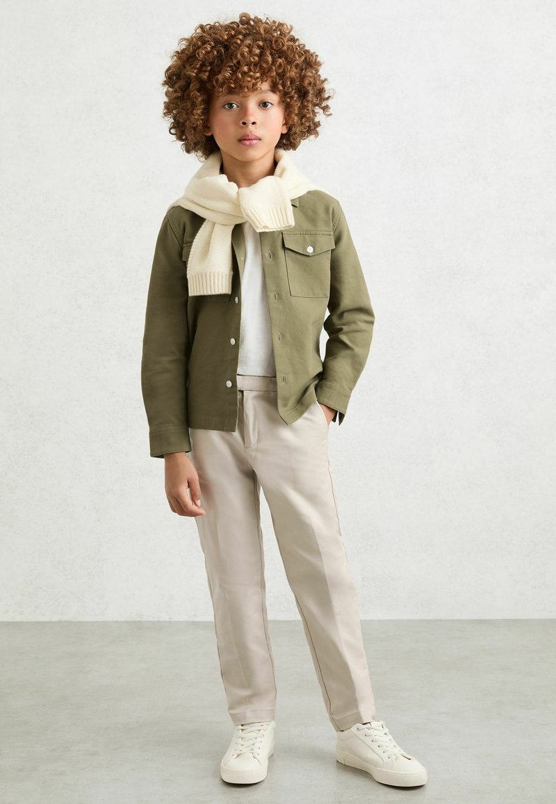 Enfant aux cheveux bouclés portant une veste vert olive, une chemise blanche, un pantalon beige, des baskets blanches, et une écharpe crème drapée sur les épaules.