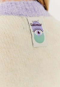 Krémový pletený svetr s měkkou texturou, s lehce fialovým žebrovaným límcem a štítkem, který obsahuje grafiku oka a slovo "Pleteniny".