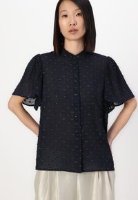 Blusa blu navale con maniche corte a sbuffo, a pois texturizzati e chiusura frontale con bottoni. Abbinata a pantaloni leggeri, lucidi e plissettati.