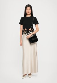 Black cut-out top with floral design, paired with a silky beige maxi skirt. Model holds a small black handbag and wears cream pointed shoes.
