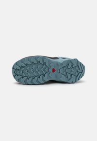 Trail running shoe sole featuring a rugged design in light blue and black, textured with multi-directional treads and a red accent.