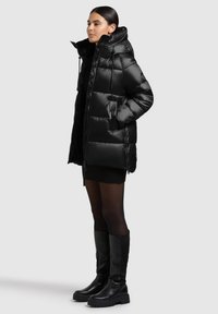 Czarna pikowana kurtka puchowa z błyszczącym, przeszywanym zewnętrzem, kapturem i kieszeniami na zamek; modelka ma na sobie czarną dzianinową sukienkę i czarne kozaki do kolan.