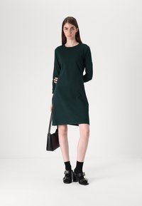 Σκούρο πράσινο φόρεμα με μακριά μανίκια, φτιαγμένο από μαλακό ύφασμα, με στρογγυλή λαιμόκοψη και χαλαρή εφαρμογή, σε συνδυασμό με μαύρα παπούτσια και τσάντα.