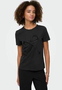 Kvinde med krøllet hår iført en sort t-shirt med et tekstureret snoet bånd-design, parret med sorte bukser og guldarmbånd.