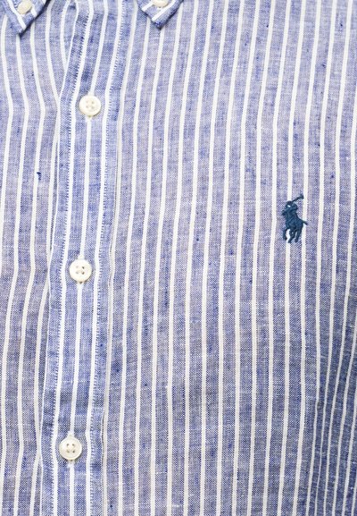 Μπλε και λευκό κατακόρυφα ριγέ πουκάμισο με λευκά κουμπιά και ένα μικρό κεντημένο λογότυπο σκορπιστή σκοπέλου σε σκούρο μπλε.