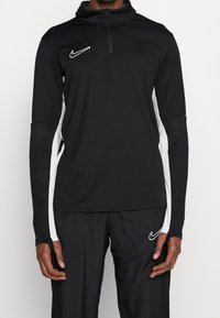 Black athletic long-sleeve shirt with a quarter-zip, white side stripes, and a small white Nike logo on the chest. Textured, moisture-wicking fabric.