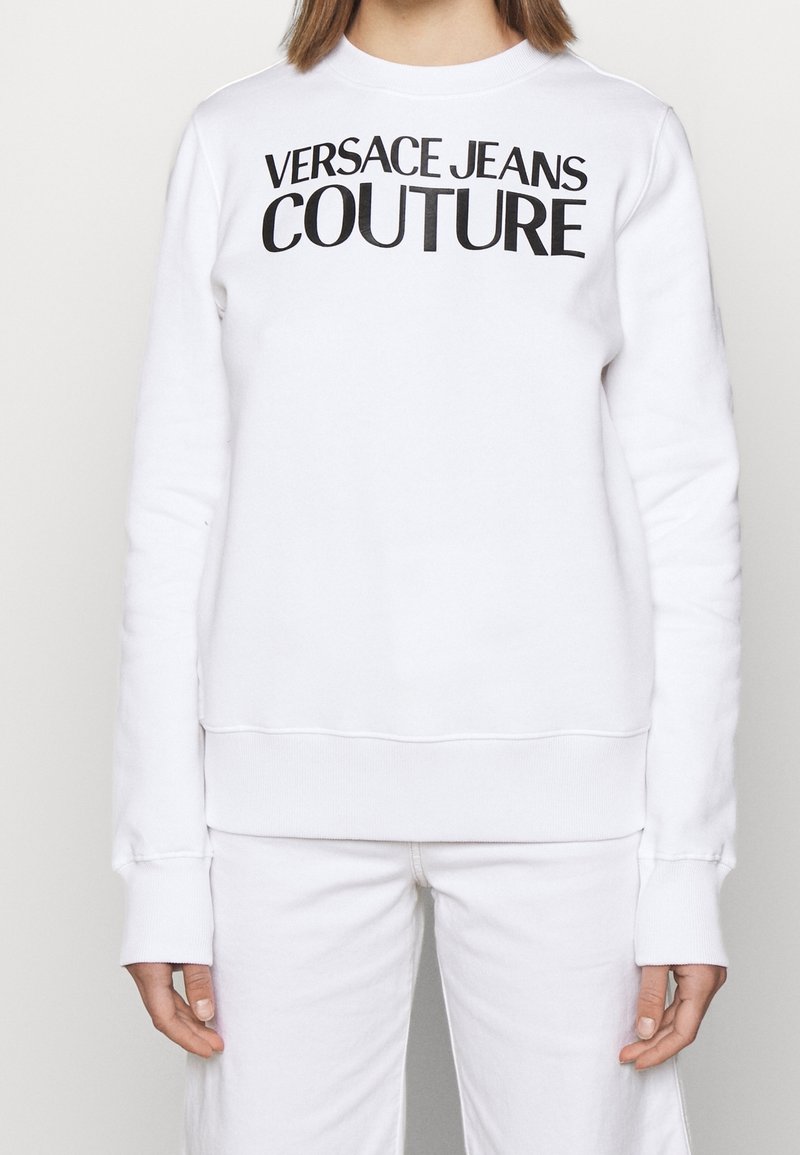 Bílá mikina s kulatým výstřihem, dlouhými rukávy, ribbovanými manžetami a lemem, a černým logem "VERSACE JEANS COUTURE" na přední straně.