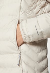 Lys beige dunjakke med diagonalt quiltet mønster; har Omni-Heat merke på mansjetten og glidelåslomme på siden.