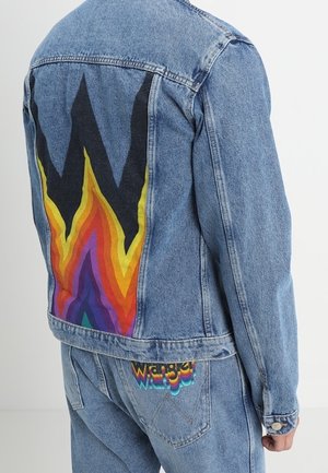 Chaqueta vaquera con un gran gráfico de llamas multicolor en la espalda, combinada con jeans vaqueros a juego que presentan un colorido logo de "Wrangler" en el bolsillo.