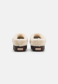 Bruna slip-on skor med en mjuk krämfärgad inredning, rundad form och en kontrasterande röd och vit "HUNTER"-logotyp på baksidan.