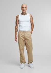 Beige chino bukser med lige pasform, høj talje og forlommer. Båret med en hvid tanktop og hvide sneakers, på en grå baggrund.