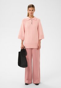 Žena v světle růžovém pleteném svetr s límcem na knoflíky a odpovídajícími širokými kalhotami, držící velkou černou kabelku.
