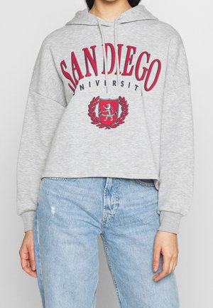 Person i en grå kort hoodie med "San Diego University" och ett rött sköld, matchat med ljusblå jeans med hög midja.