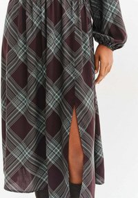 Długa bordowa sukienka w kratę w kolorach czarnym, białym i jasnoniebieskim. Posiada długie rękawy oraz rozcięcie po boku. Miękka tekstura tkaniny.