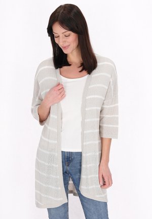 Frau mit dunklen Haaren, die eine hellgrau-weiß gestreifte Strickjacke, ein weißes Top und blaue Jeans trägt, schaut mit einem leichten Lächeln nach unten.
