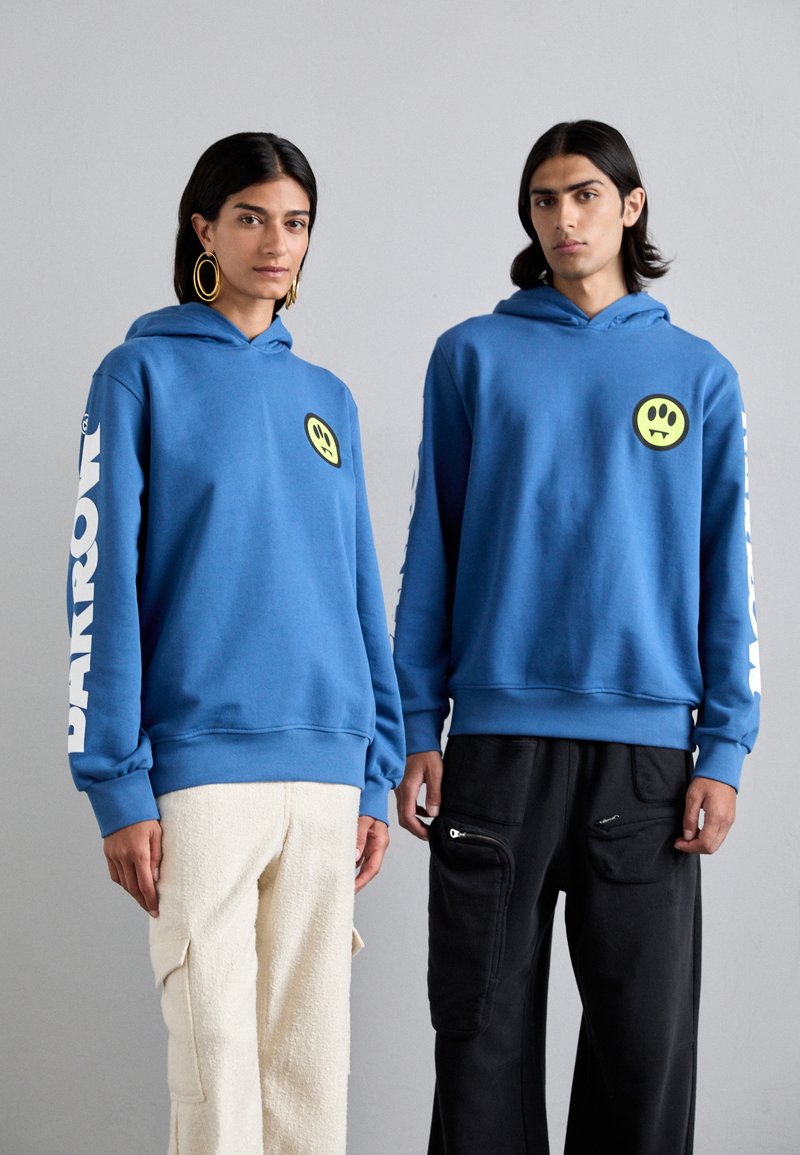 Blå hetter med gule smiley-ansiktslapper; en har 'BARROW' i hvitt på ermet. Kombinert med beige fluffy og svarte glidelåsbukser.