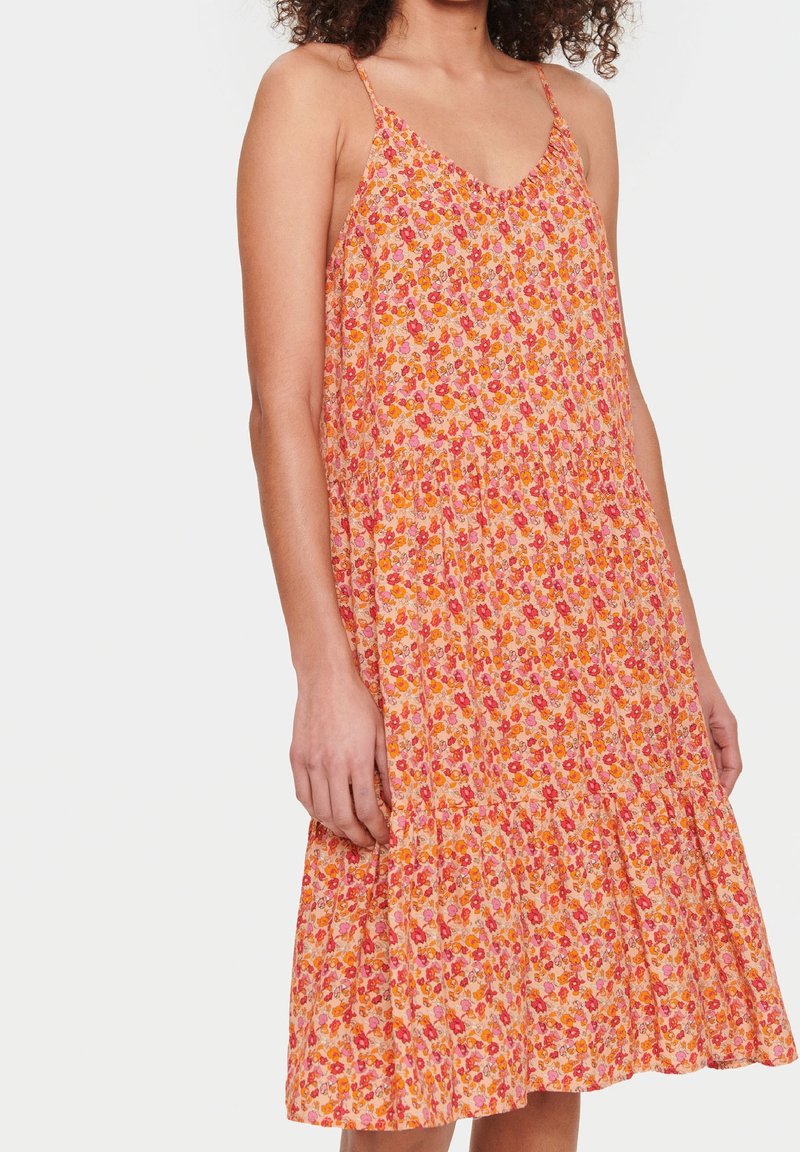 Femme portant une robe midi sans manches à volants avec fines bretelles et motif floral rose, orange et rouge, debout sur fond blanc.