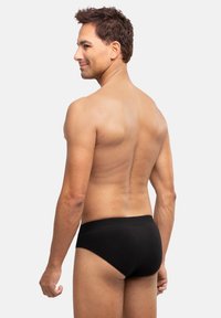 Schwarze Baumwollunterhose, schlichtes Design. Elastischer Bund, eng anliegende Form. Modell von hinten gezeigt mit kurzem dunklem Haar, neutraler Hintergrund.