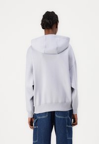 Sudadera con capucha gris claro de corte relajado, con hombros caídos, puños y dobladillo ribeteados. Textura suave, sin patrones ni herrajes visibles.