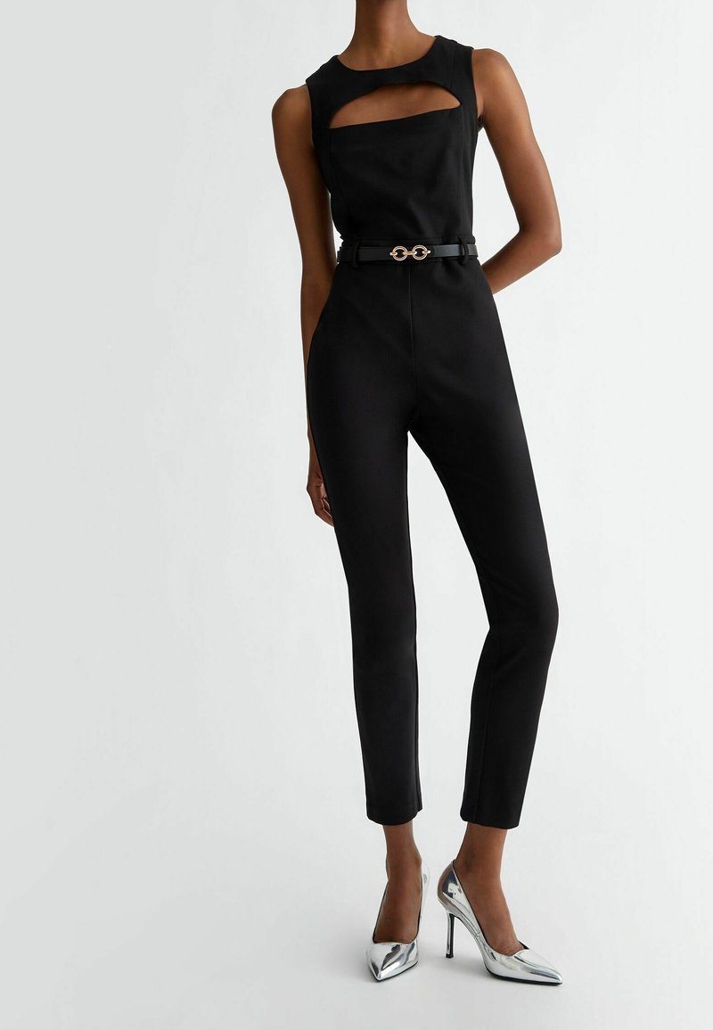 Femme portant une combinaison noire sans manches avec une découpe poitrine et une ceinture noire, associée à des escarpins argentés à bout pointu.