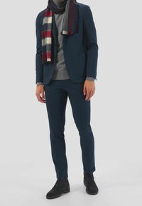 Abito blu navy con pantaloni a sigaretta e blazer coordinato. Dolcevita grigio indossato sotto, accompagnato da una sciarpa a quadri in rosso, blu navy, bianco e grigio. Stivaletti neri completano il look.