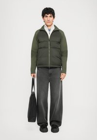 Olive green padded jacket with a zip closure, grey wide-leg jeans, and black shoes. Black tote bag in hand, standing against a white background.