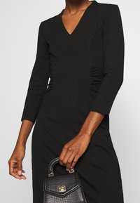 Robe noire avec un décolleté en V et des manches trois-quarts, présentant des détails froncés à la taille et un ajustement slim. Sac à main noir texturé porté à la main.