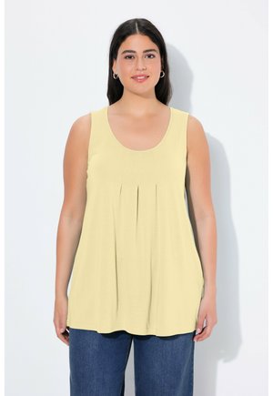 Young woman with long dark hair wears a pale yellow sleeveless top and dark blue jeans, standing against a plain white background.