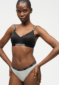 Femme aux cheveux tressés portant un soutien-gorge noir Calvin Klein et un sous-vêtement gris Calvin Klein, debout les mains sur les hanches devant un fond uni.