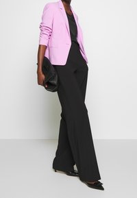 Světle růžový blejzr s vypasovaným střihem, černý top, černé kalhoty s širokými nohavicemi a černé lodičky s špičatou špičkou, držící černou psaníčko.