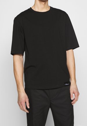 Mann trägt schlichtes schwarzes Kurzarm-T-Shirt und schwarze Hose, steht vor einem einfachen hellgrauen Hintergrund.