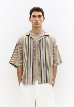 Kortærmet skjorte i beige og blå striber, med knaplukning og afslappet pasform. Båret over en grå T-shirt, parret med hvide bukser.