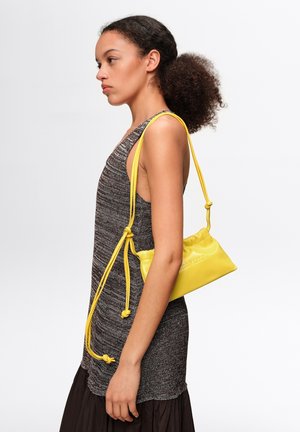 Mujer con un vestido sin mangas y textura, llevando un pequeño bolso amarillo con correas anudadas sobre el hombro, de perfil.