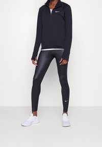 Czarny sportowy top z długim rękawem, z zamkiem błyskawicznym do ćwierć, z otworami na kciuki, zestawiony z błyszczącymi czarnymi legginsami i białymi butami sportowymi. Obecne logo Nike.