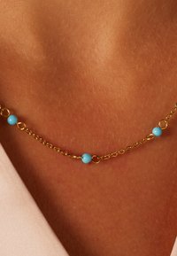 Gouden ketting met afwisselend lichtblauwe kralen, delicate schakels en een gladde afwerking, die tegen de huid rust met een zachte textuur.