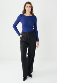 Koszula w niebieskim kolorze z długim rękawem i prążkowaną teksturą, zestawiona z ciemnoszarymi spodniami o prostym kroju i wysokim stanem. Modelka stoi z rękami w kieszeniach.