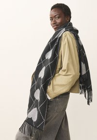 Black and grey patterned scarf featuring large hearts and diamond shapes, fringed edges, worn over a light khaki jacket.