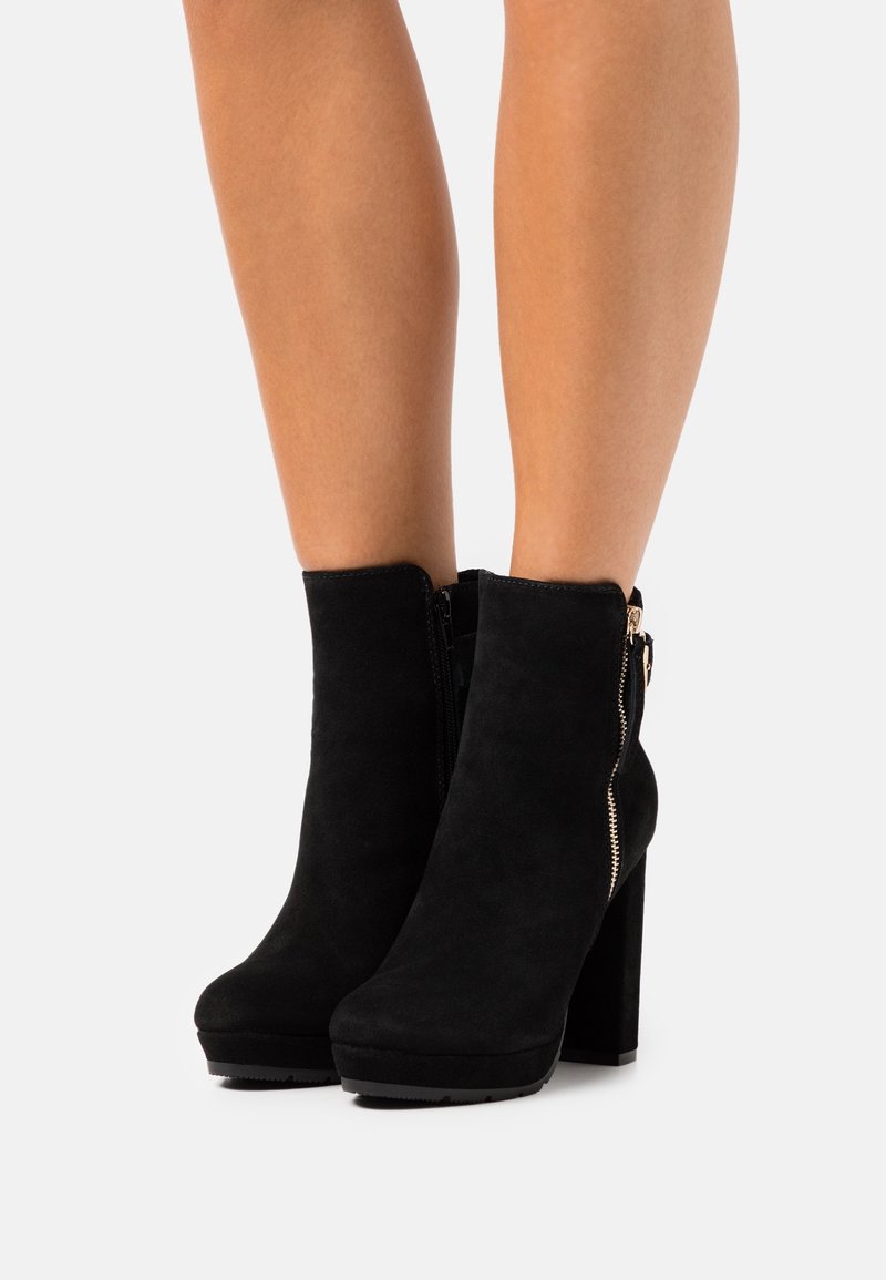 Jambes portant des bottines noires en daim à talons hauts avec une fermeture éclair latérale sur un fond uni.