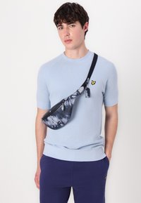 Svetle modrý svetr s krátkým rukávem a texturovaným vzorem, spárovaný s tmavě modrou taškou přes rameno s tie-dye vzorem, která má zipové uzavření a boční detaily.
